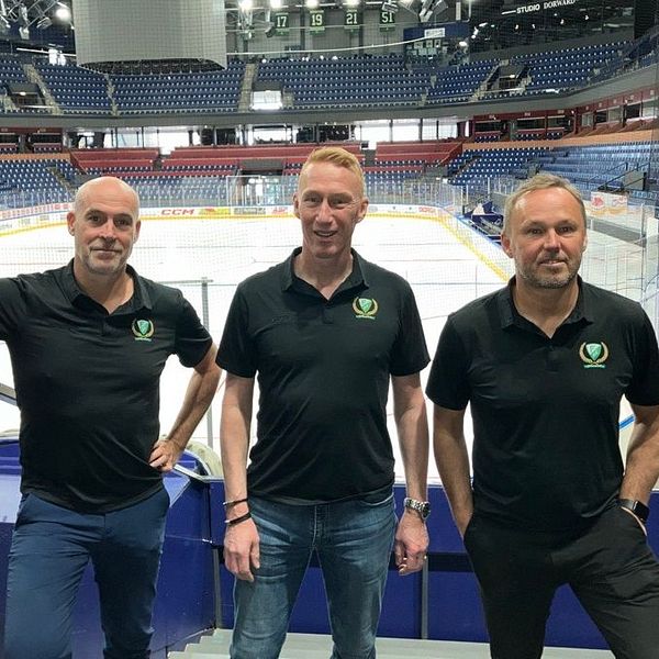 Tre personer i svarta pikétröjor står vid läktaren i en tom ishockeyarena.