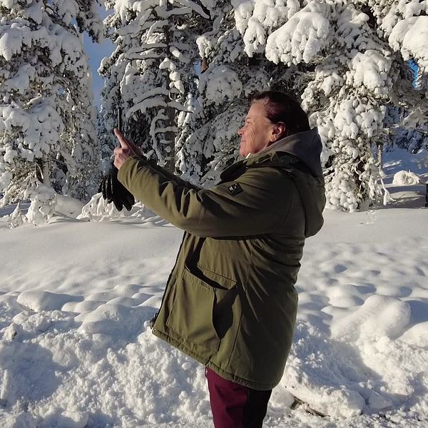En medelålderskvinna står i grön jacka i ett snölandskap, hon står vänd mot solen och tar en bild med mobiltelefonen.