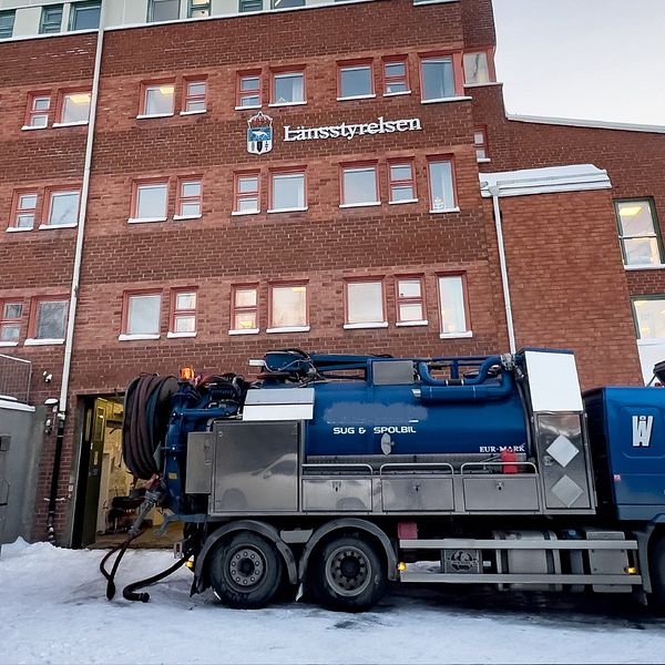 En snöklädd gård framför en tegelbyggnad med texten ”Länsstyrelsen” synlig. En pumpbil står utanför och pumpar vatten.