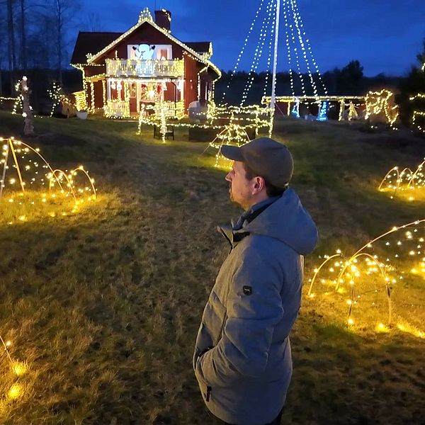 Kristoffer Rudin står på en grön gräsmatta som är täckt av strålande julbelysning. I bakgrunden syns ett rött hus dekorerat med flera ljus, och trädgården är prydd med lysande former i olika konfigurationer, inklusive bågar och spiraler. Stämningen är festlig och mysig, med en mörkblå himmel som ger en vacker kontrast till det gyllene ljuset.