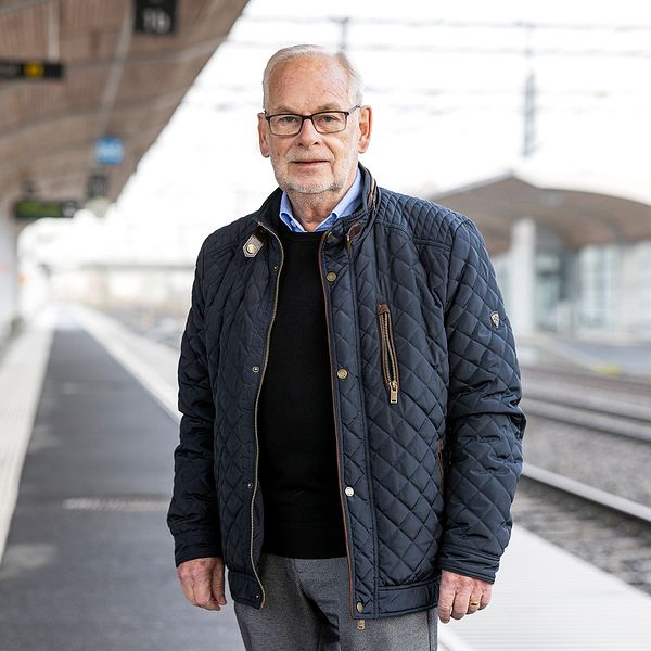 En person står på en tom tågplattform klädd i en mörk jacka och grå byxor. Plattformen är delvis skyddad av ett tak, och tågspåren sträcker sig bort i bakgrunden.