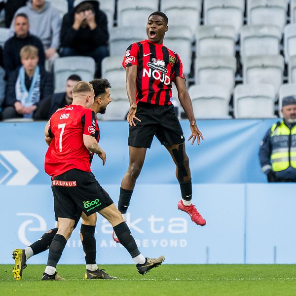 Två fotbollsspelare i rödsvarta tröjor firar mål under en match inför publik.