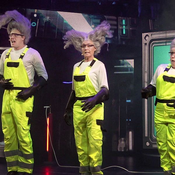 Tre personer i neonfärgade arbetskläder med reflexdetaljer står på en scen. De har alla liknande frisyrer i form av stora yviga gråa hår. Bakgrunden visar en teknologisk miljö med futuristiska inslag.