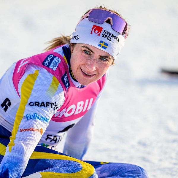 En tävlingsåkare i längdskidåkning utomhus, klädd i färgglad tävlingsdräkt med synliga sponsornamn, sitter på knä i snön.