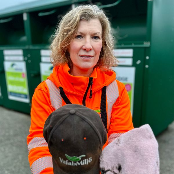 En person i en orange arbetsjacka håller en svart keps med en logotyp och en grå handduk, med en avfallscontainer i bakgrunden.