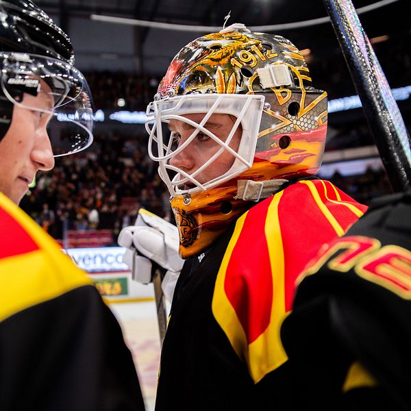 En grupp ishockeyspelare, med fokus på en målvakts hjälm som har en färgglad design med inslag av röd och gul. Spelarna bär matchtröjor i liknande färger, vilket skapar en enhetlig och dynamisk atmosfär. I bakgrunden syns en publik som är engagerad i matchen.