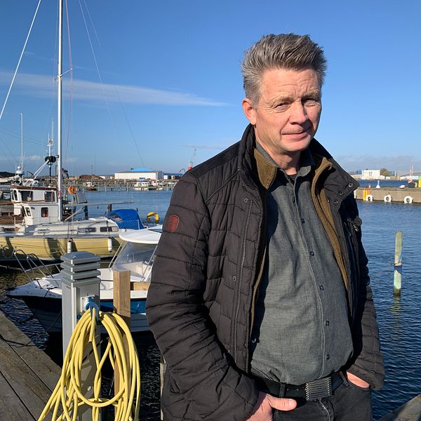 En person står på en brygga vid en hamn, omgiven av segelbåtar och en klar blå himmel. Bryggan är gjord av trä, och det finns en sladd av gult material som hänger i förgrunden. I bakgrunden syns byggnader och kranar som bidrar till en marin atmosfär.