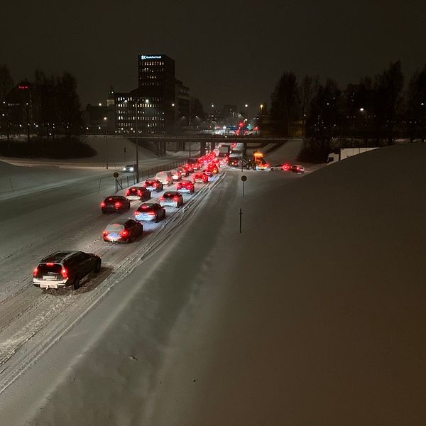Stadsmiljö med bilkö i snöigt väder under natten. En rad bilar syns körande i det begränsade ljuset från gatlampor och bilars bakljus. På vägarna finns snö som skapar hinder. I bakgrunden syns målningsliknande byggnader belysta av stadens ljus.