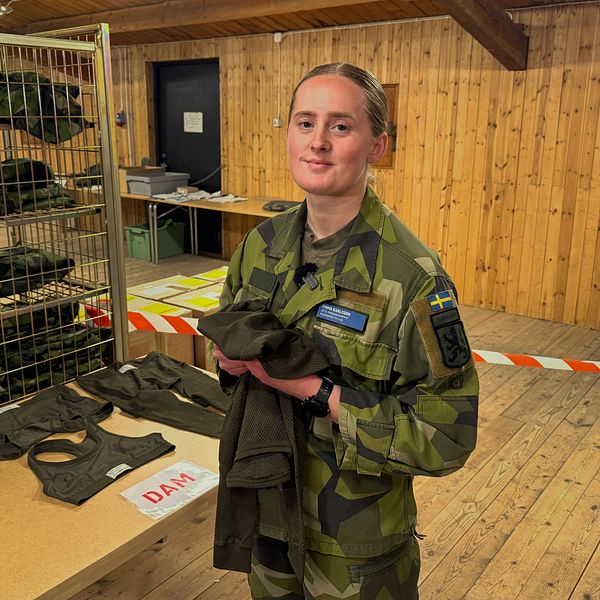kvinna i gröna uniformskläder står inomhus och håller i en underställströja vid ett bord märkt ”DAM”