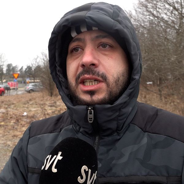 En person står utomhus i vinterkläder och håller en mikrofon med ”SVT” tryckt på, i förgrunden. I bakgrunden syns två personer och flera parkerade bilar längs en väg med träd utan löv.