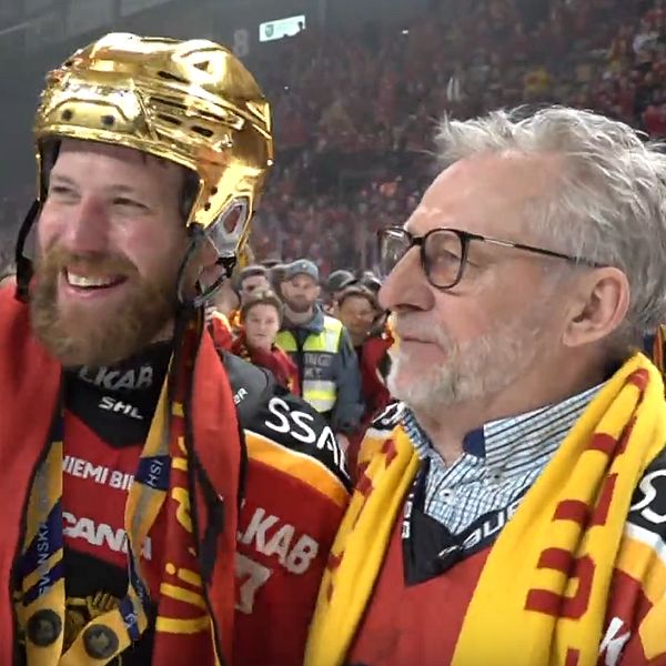 Två personer i hockeytröjor firar på isen efter match, omgivna av publik och funktionärer.