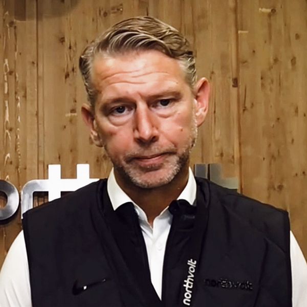 En person står framför en träpanel som har en logotyp för ”nothnet”. Personen bär en svart väst över en vit skjorta. Bakgrunden är mörk och varm, vilket skapar en professionell atmosfär.