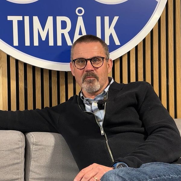 Person sitter avslappnat i soffa framför Timrå IK:s stora klubbmärke på väggen.