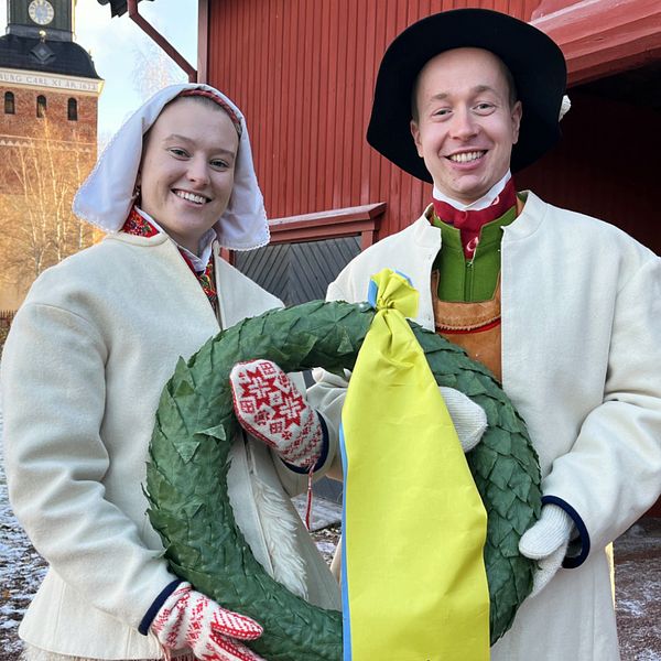 Bild på kransparet Elsa och Gustav framför Mora kyrka, klädda i dräkt och med en krans i händerna.