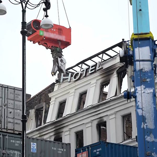 En kran som lyfter en röd kapsel över ett byggnadsområde, med en del av en byggnad i bakgrunden där skylten ”HOTEL” är synlig. Området är omgivet av fraktcontainrar.