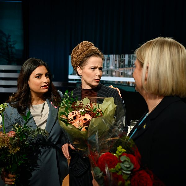 Tre personer står på en scen och håller blombuketter vid ett evenemang eller prisutdelning.