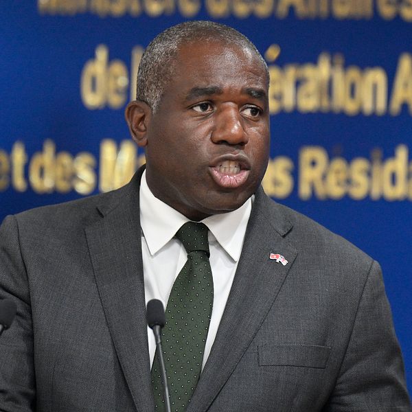 Man i kostym talar vid podie med brittisk flagga och blå bakgrund med gul text.