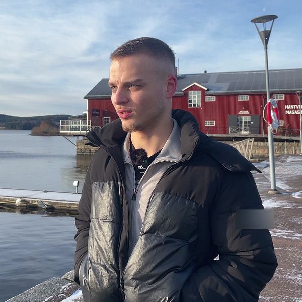 En person står på en brygga vid en sjö, klädd i en svart jacka. I bakgrunden syns ett rött hus och en lugn vattenyta med bergen i fjärran. Det är en klar dag med några moln på himlen.