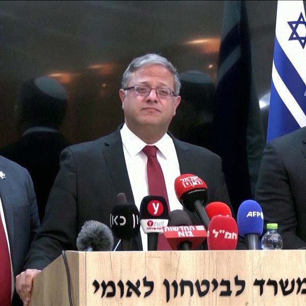 Tre personer med kippa står vid ett podium med mikrofoner och medieutrustning under en presskonferens. I bakgrunden syns en israelisk flagga och en skylt med hebreisk text, vilket indikerar att evenemanget äger rum i Israel. Personerna är formellt klädda i kostymer med en av dem i röd slips.