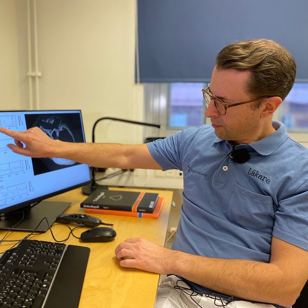 En läkare sitter vid ett skrivbord och pekar mot en datorskärm där en bild på en EKG-kurva syns.
