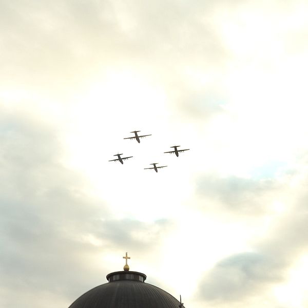 Fyra flygplan passerar över en kupolformad byggnad under en molnig himmel. En gyllene kors syns på toppen av kupolen.