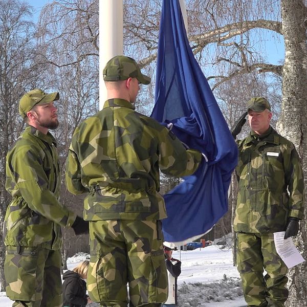 Tre militärer står vid en flaggstång där Natoflaggan håller på att hissas