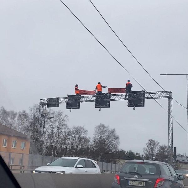 Tre personer med banderoll på stålbalk ovanför motorvägen