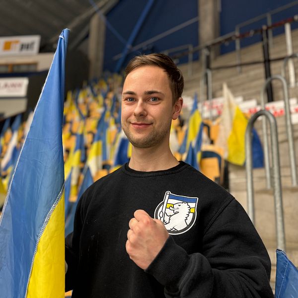 Person i svart tröja med klubbemblem håller blå-gul flagga på läktaren.