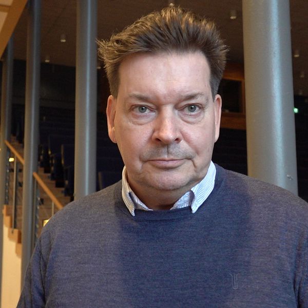 En person iklädd en mörk tröja står i en nästan tom föreläsningssal. I bakgrunden syns en trappa och rader av tomma stolar.