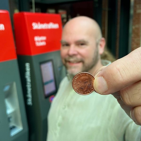 En person håller upp ett mynt framför två biljettautomater från Skånetrafiken. Automaterna har tydlig information om biljettköp på svenska och engelska.