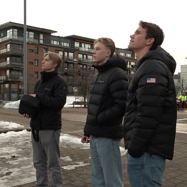 Tre personer står på en trottoar med moderna byggnader i bakgrunden. De bär svarta jackor och verkar titta upp mot himlen. Snö och is syns på marken i några områden.