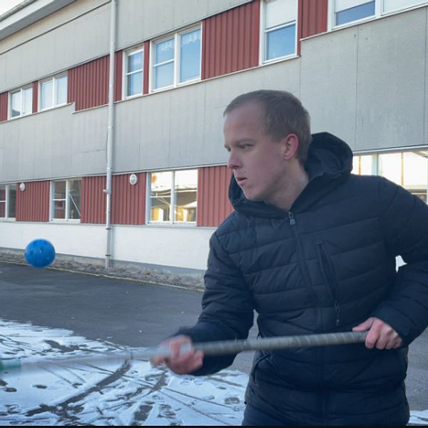 En person som svingar en sportutrustning i en skolgård, med en byggnad i bakgrunden. Snö täcker en del av marken, och flera fönster syns på byggnaden. En blå boll och en ramp för skateboarding eller liknande aktivitet är synliga i scenen.