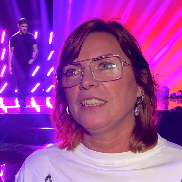 Susanne Bertlin, sångpedagog, står framför en scen med starka ljuseffekter och en manlig artist som uppträder i bakgrunden. Scenen har ljusa, färgglada strålkastare som skapar en livlig atmosfär.