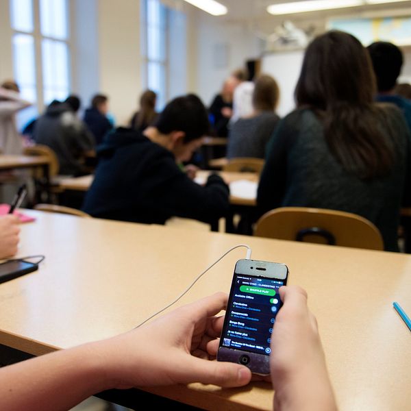 En student sitter på en bänk i ett klassrum och håller en smartphone som visar en musikapp. I bakgrunden syns andra elever som är upptagna med skrivande eller antecknande. Rummet har stora fönster som släpper in naturligt ljus.