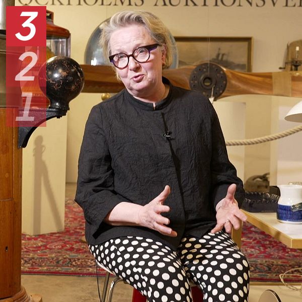 Designexperten Anna-Kaisa Öhgren sitter bredvid maritim utrustning och berättar, bord med porslin i förgrunden.