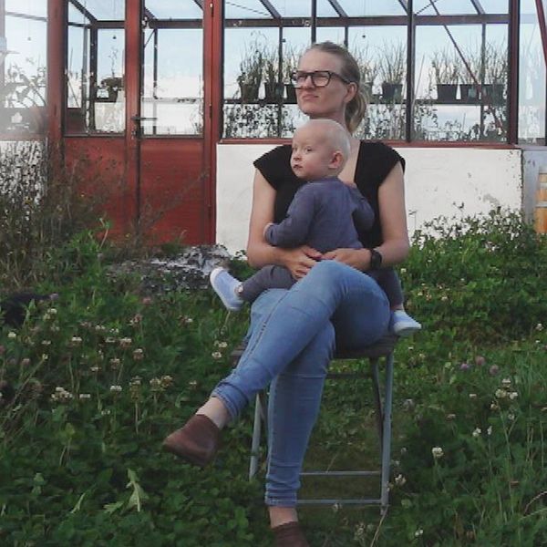 Petra Eklund och hennes cirka 1-åriga son sitter på en stol i en grönskande trädgård framför ett växthus