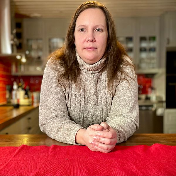 En person i en grå tröja sitter vid ett matbord med en röd duk, omgiven av en hemtrevlig köksmiljö med träelement och bakgrundsdetaljer som köksredskap och hyllor.