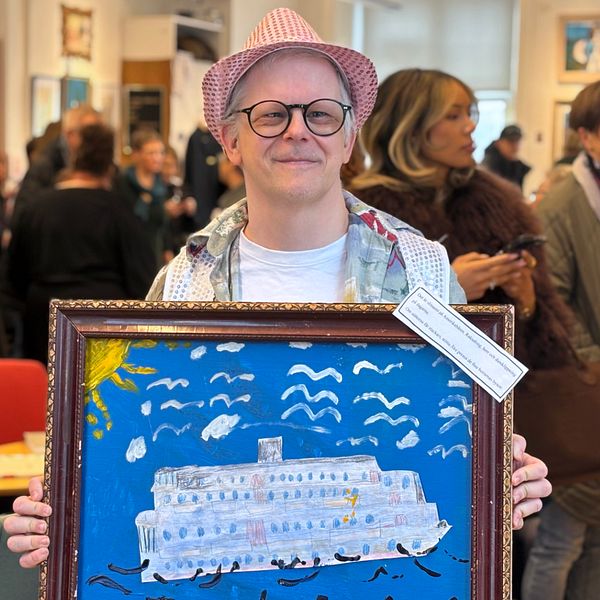 Kristoffer Gullersbo håller i sin tavla som visar ett fartyg omgiven av måsar på en blå bakgrund. I bakgrunden syns flera människor i ett rum, som verkar vara på en utställning eller liknande evenemang.