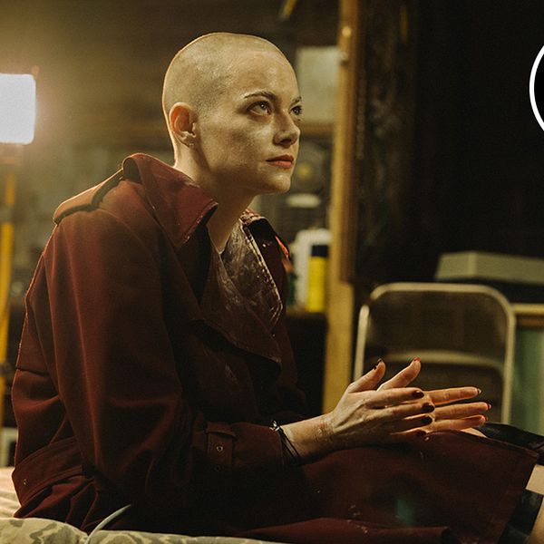 Emma Stone sitter på en säng i en mörk, rörig miljö i filmen Bugonia.