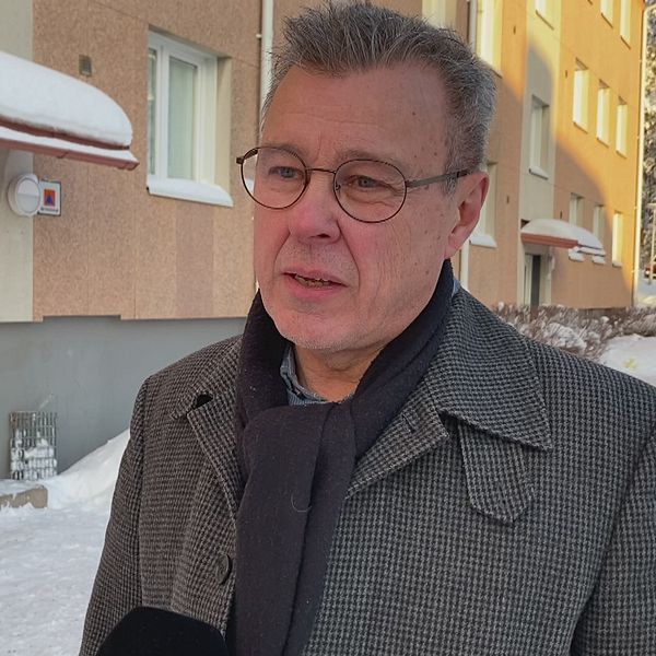 En man klädd i en mörk kostymjacka står utomhus framför en flerfamiljshus täckt av snö. I bakgrunden syns en entrédörr och snötäckta bilar längs gatan som skapar en vinterlig atmosfär.