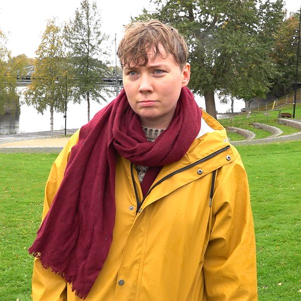 Ellen Ström står på en grön gräsmatta med en gul regnjacka och en röd scarf. I bakgrunden syns Umeåälven med spegelblanka ytor och omgivande träd, vilket ger en höstlig atmosfär. Det är en lugn och svagt molnig dag.