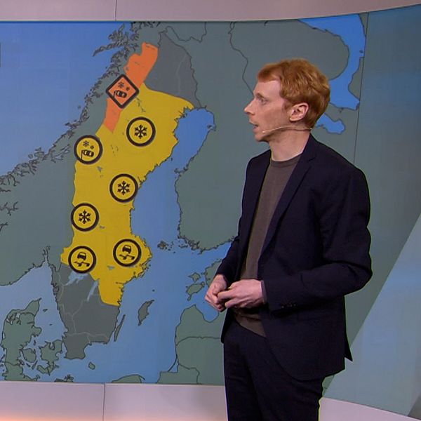 En person står framför en väderkarta som visar varningar för olika väderförhållanden i Sverige. Kartan har områden markerade i orange och gult som indikerar varningar, tillsammans med symboler för snö och andra möjliga faror. Texten ”Varningar” är synlig i övre vänstra hörnet av kartan.
