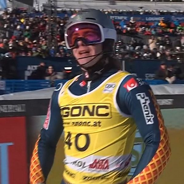 En skidåkare i en gul outfit med nummer 40 på bröstet går längs en bana, omgiven av en stor publik som står på läktarna. Soligt väder och en klar blå himmel skapar en festlig atmosfär kring tävlingen.