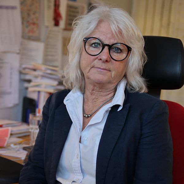 En person sitter vid ett skrivbord i ett kontorslandskap, omgiven av högar av papper och kontorsmaterial. På skrivbordet syns en dator med en öppen skärm och diverse föremål som en mobiltelefon och en penna. Bakgrunden är fylld med dokument och notiser som hänger på väggen.