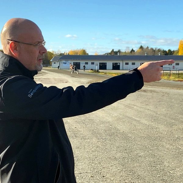 En man står på en grusplan och pekar framåt med höger hand. I bakgrunden syns byggnader och en travghäst med ett höstigt landskap. Himlen är klar med några få moln.