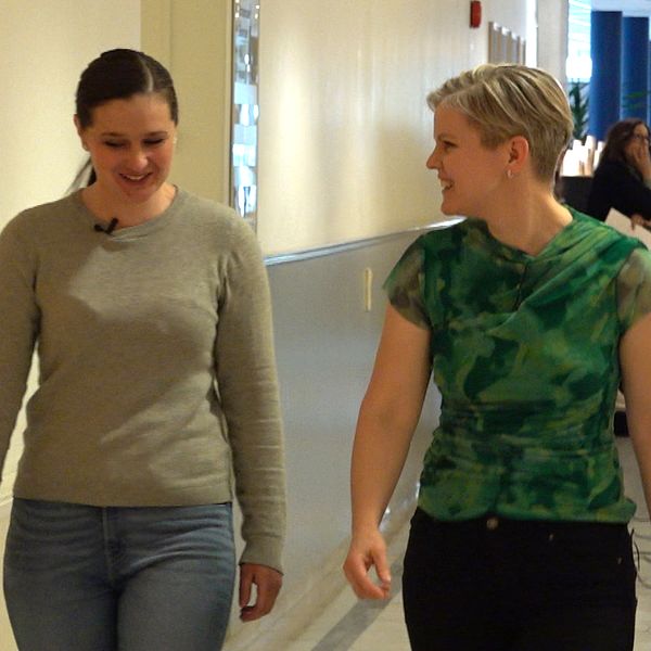 Två personer promenerar i en skol- eller vårdkorridor med fler människor i bakgrunden.