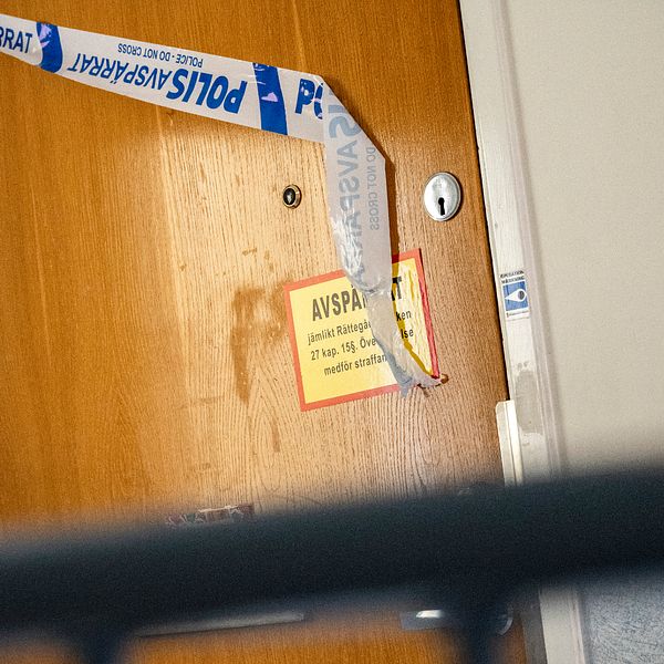 En stängd dörr märkt med polisens avspärrningsband och en gul skylt med texten ”AVSPÄRRAT”. Dörren visar tecken på slitage med fläckar synliga på ytan. Misstänkt mord i Lund.