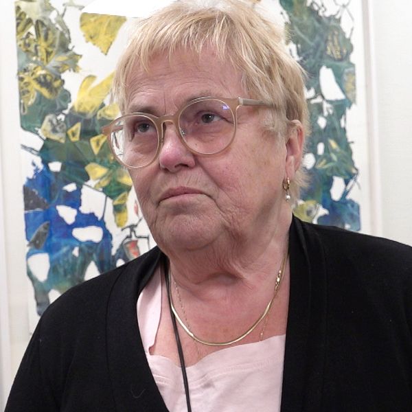 En äldre kvinna med ljust hår står framför en abstrakt tavla på en vit vägg.