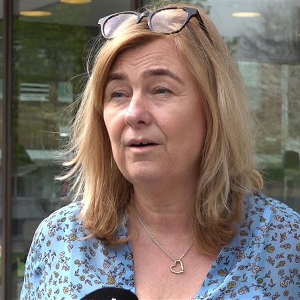 Kvinna med blont hår och blåmönstrad skjorta intervjuas utomhus framför glasdörrar.