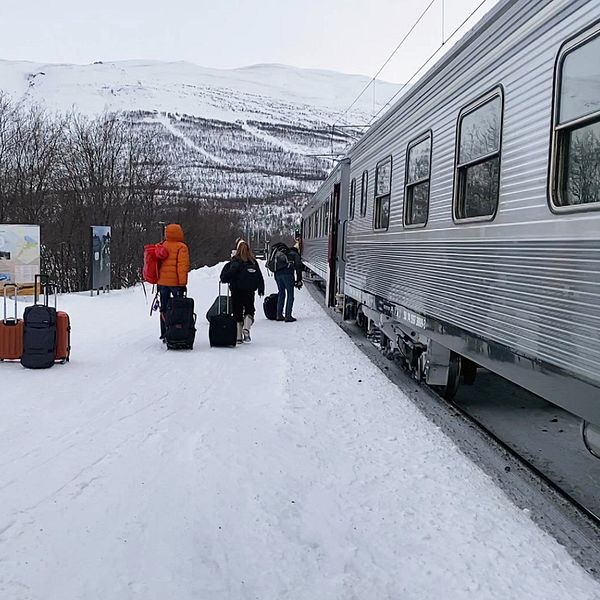 Passagerare väntar på en tågstation i ett snöigt landskap, med en tågvagn synlig till höger. Flera personer är klädda i vinterkläder och har bagage. I bakgrunden syns snötäckta fjäll och en informationsskylt vid stationen.